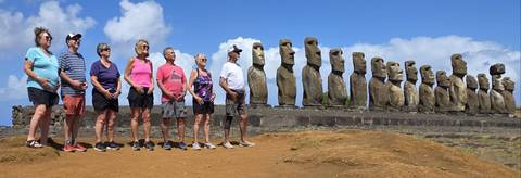       Visitors stand in line mirroring a long row of moai under a blue sky.
  