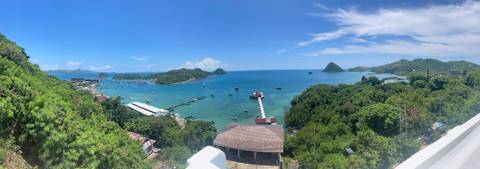       Wide coastal panorama of Labuan Bajo harbour, scattered boats and green islets under clear skies.
  