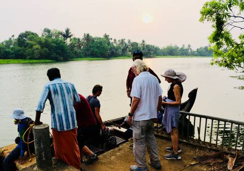       Travelers board a small wooden ferry on a tranquil river as the sun sets through haze.
  