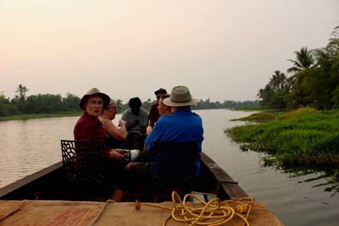       Elderly tourists sit in an open motorised canoe gliding along Kerala backwaters lined with greenery.
  