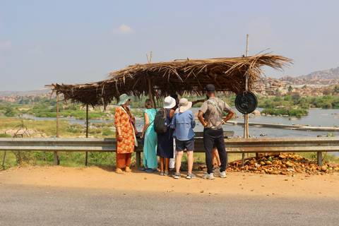       Travelers stand under a thatch shelter beside a river valley dotted with boulders and palms.
  