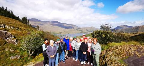       Tour group posing on a scenic overlook above a lake and mountains in Ireland under partly cloudy sky.
  
