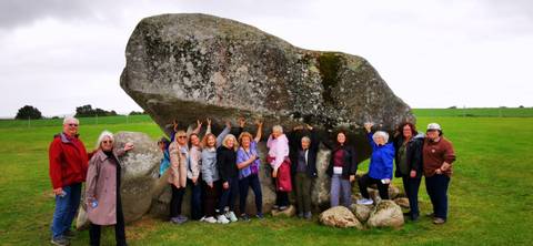       Large group standing under and supporting a massive glacial erratic boulder in a green Irish field.
  