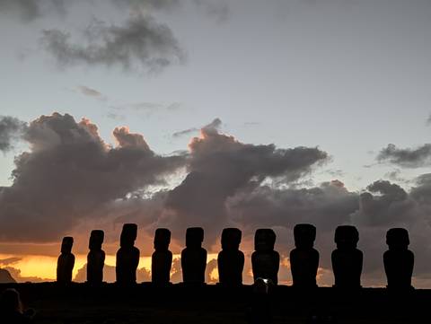       Silhouetted row of Easter Island moai statues stands beneath colourful twilight clouds.
  
