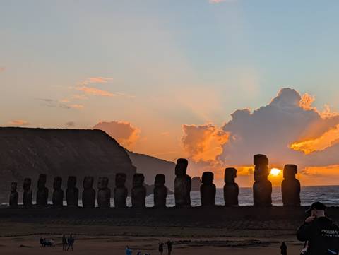       Sun rises between moai statues lined along the coast, casting rays through dramatic clouds.
  