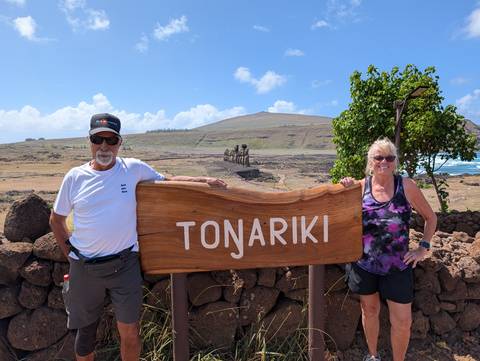      An older couple poses beside a wooden Tongariki sign with the iconic Easter Island moai statues and arid hills in the background under a bright blue sky.
  