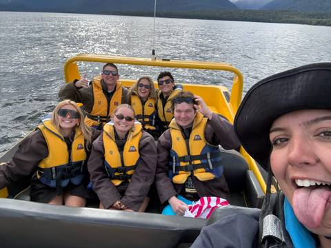       Smiling friends in yellow life jackets sit in a speedy yellow jet boat on a lake taking a selfie.
  