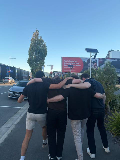       Five young men walk arm-in-arm down a modern city street as evening light falls.
  