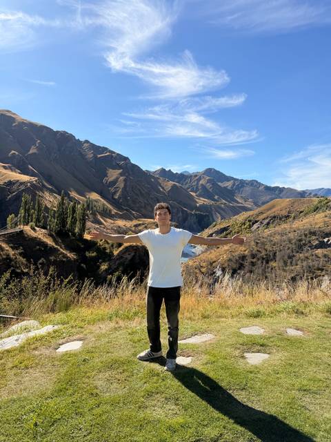      A young man in a white T-shirt spreads his arms wide before a dramatic New Zealand river gorge and mountains.
  
