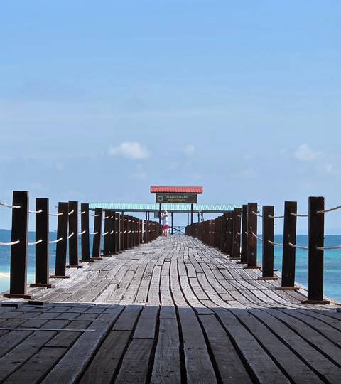       A long wooden jetty stretches over turquoise sea toward a small shelter, with a lone person standing at the far end.
  