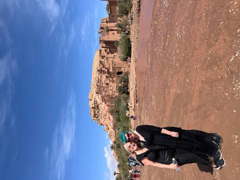       Two women stand smiling before the earthen kasbah of Aït Ben Haddou under a blue sky.
  