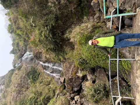       Smiling tourist posing at a railing in front of a tall cascading waterfall on a lush hillside.
  