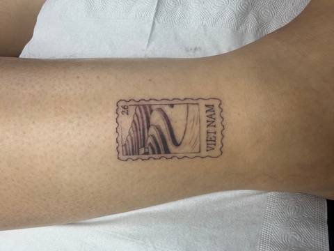      Close-up of a small postage-stamp style tattoo reading 'VIET NAM' with a winding road illustration on a person's leg.
  