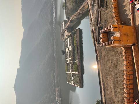       Elevated view of Amber Fort's symmetrical garden island reflected in a tranquil lake with hazy sunlit hills beyond.
  