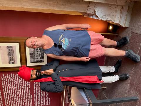       Tourist in casual attire standing next to a traditionally dressed Indian host with red turban inside a historic building.
  