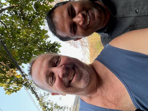       Close selfie of two smiling men outdoors beneath leafy tree branches.
  