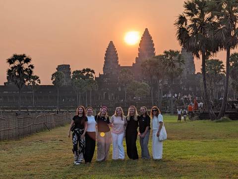       Group of friends posing at sunrise in front of Angkor Wat’s iconic towers and palm trees.
  