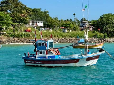       Colorful fishing boat named 'Deutsch Lanka' anchored on turquoise water with coastal village behind.
  
