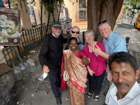       Travellers and a local woman smile for a street-side selfie beside a tree and iron fence.
  