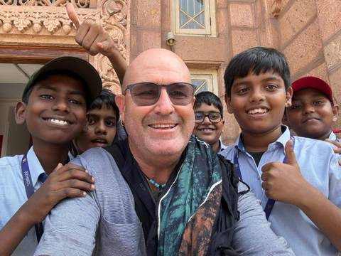       Middle-aged traveller poses with cheerful schoolboys giving thumbs up outside a sandstone building.
  