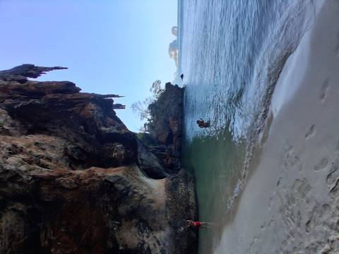       Secluded sandy cove beneath towering limestone overhang with turquoise water lapping the shore.
  