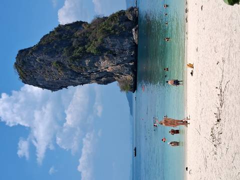       Tourists swim and relax on a tropical white-sand beach with towering limestone stack backdrop.
  