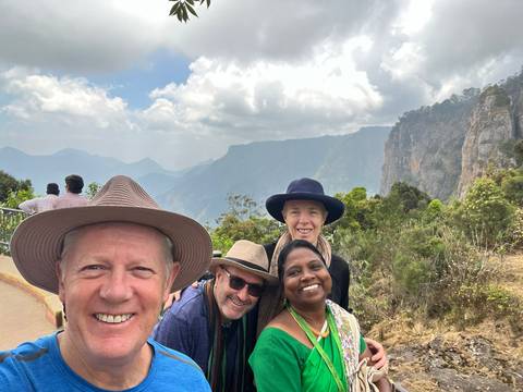       Smiling travellers pose at a misty mountain viewpoint with cliffs and deep valleys behind.
  