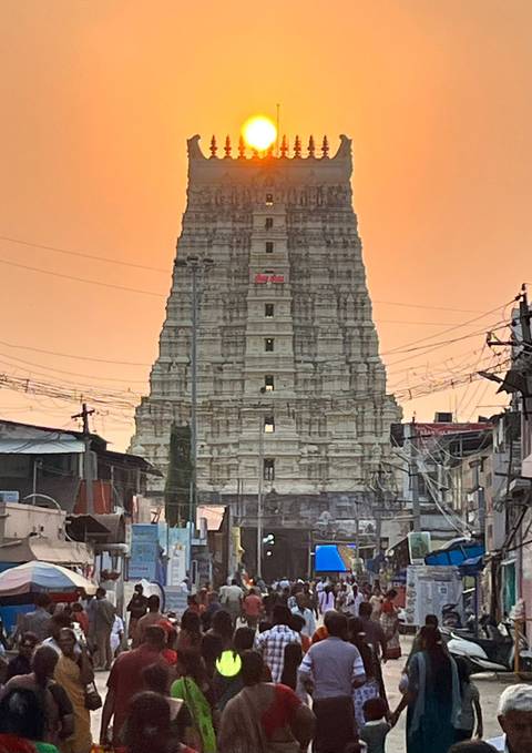       Tall gopuram temple tower rises against an orange dusk sky, framed by street wires and shops.
  