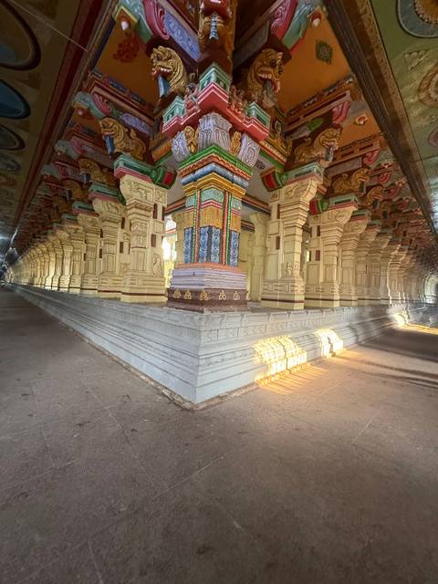       Grand ornate corridor of a South Indian temple with richly painted columns and slanting sunlight illuminating stone floors
  
