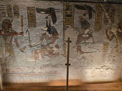       Ancient wall art showing Anubis seated with additional deities and hieroglyphs, protected by glass panels
  