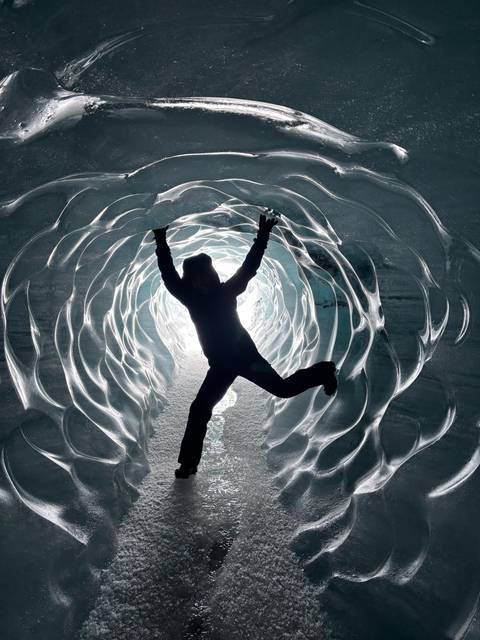       Silhouetted figure jumping inside a glistening ice cave tunnel lit from behind.
  