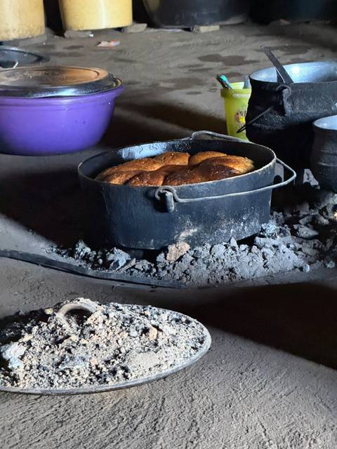       Loaf of bread baking in a black cast-iron pot on hot embers inside a rustic hut.
  