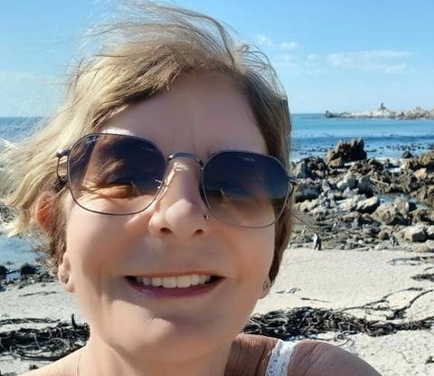       Woman taking a selfie on a rocky beach with calm blue ocean and distant penguins.
  