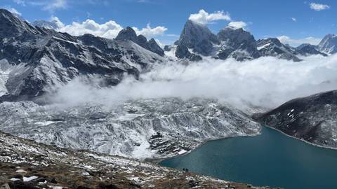       High-altitude turquoise lake surrounded by snow-dusted mountain ridges and drifting clouds.
  