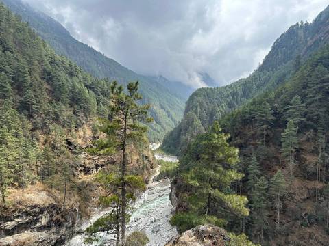       Verdant river valley with pine-covered slopes and dramatic clouds above.
  