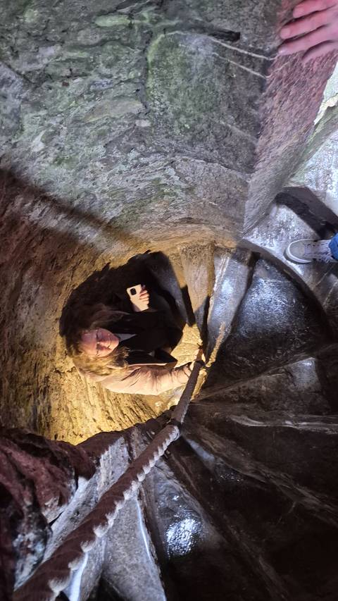       A traveller carefully descends a narrow stone spiral staircase inside an ancient tower, lit by phone light.
  