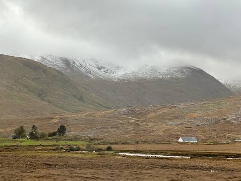       Misty, snow-dusted mountains loom over a lonely cottage in the rolling Connemara landscape.
  