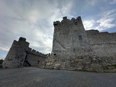       Massive grey stone fortress perches on rugged bedrock beneath a moody sky.
  