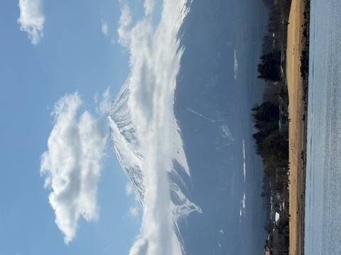       Snow-capped Mount Fuji rises above low clouds with a lake and rural foreground under a blue sky.
  