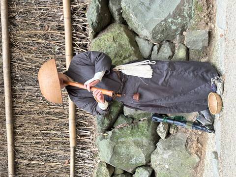       Street performer in traditional robes and straw hat plays a bamboo flute beside a stone wall.
  