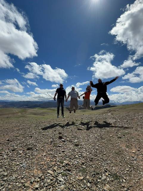       Four travellers leap simultaneously on a windy mountain plateau under bright blue skies and clouds.
  
