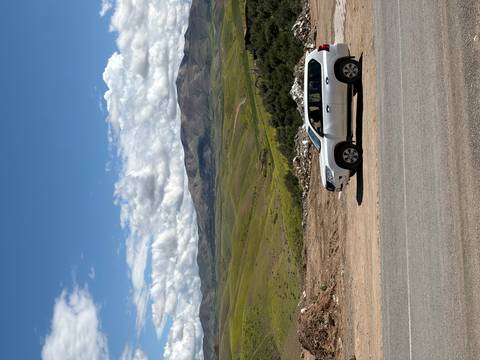       Silver 4x4 vehicle parked beside an open road with wide green valleys and rugged mountains beyond.
  