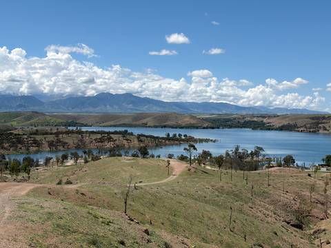       Large blue reservoir surrounded by rolling hills and distant mountain range under scattered clouds.
  