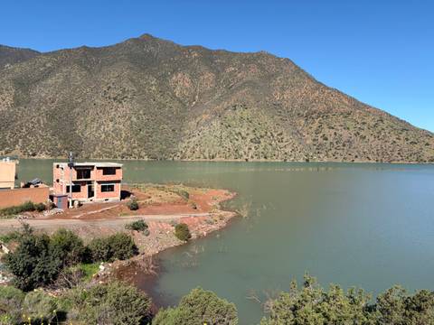       Quiet lake bordered by sloping forested mountains with a solitary unfinished brick house on the shore.
  