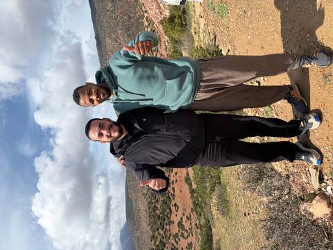       Two smiling men giving thumbs-up pose on a hilltop with patchwork valleys and clouds behind.
  