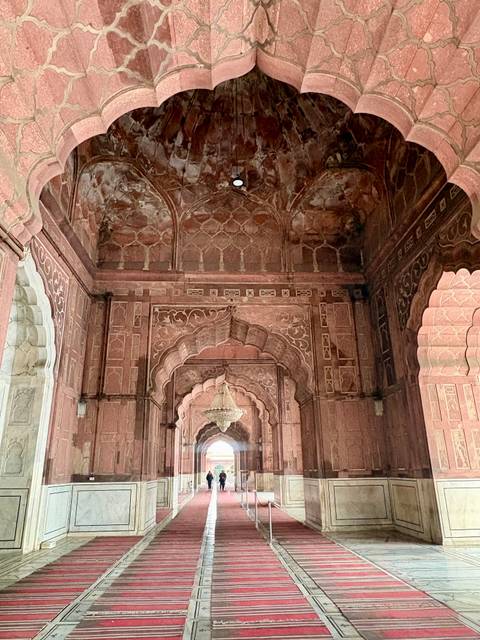       Ornate red sandstone corridor with intricate Mughal carvings and a crystal chandelier hanging above.
  