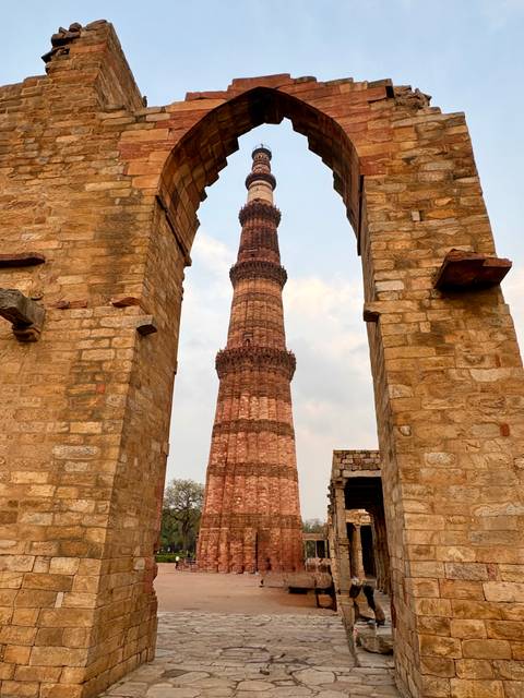       Tall Qutub Minar sandstone tower framed by crumbling stone archways under a light sky.
  