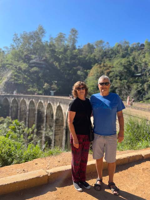       Smiling couple stands before Sri Lanka's iconic Nine Arches Railway Bridge amidst lush jungle
  