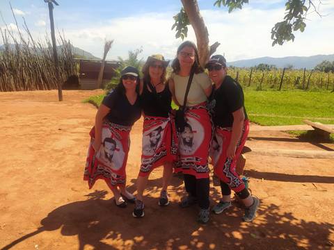       Four women wearing bright Swazi wraps pose together smiling in a sunny village setting.
  