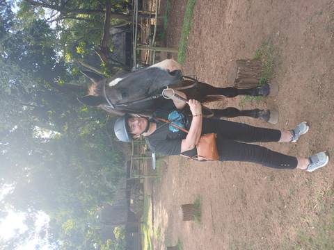      A woman wearing a riding helmet stands next to a dark horse in a shady paddock area.
  